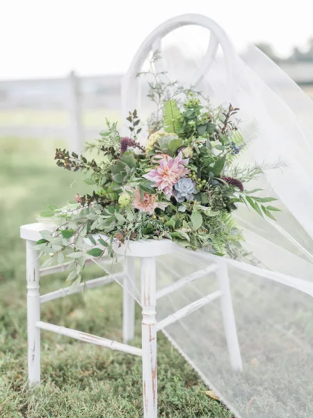 A lush floral arrangement featuring dahlias and greenery arranged on a white chair with a veil in an outdoor setting.