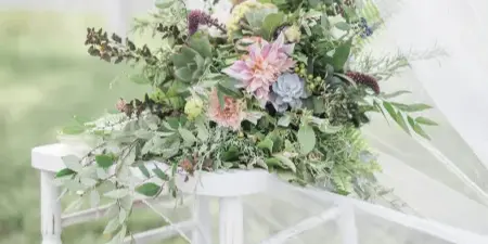 A lush floral arrangement featuring dahlias and greenery arranged on a white chair with a veil in an outdoor setting.
