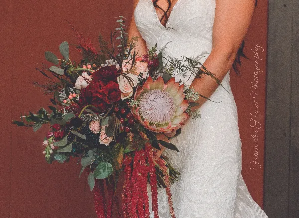 A bride in a white lace gown holding a large, custom wedding bouquet with a central protea, red roses, and trailing greenery.