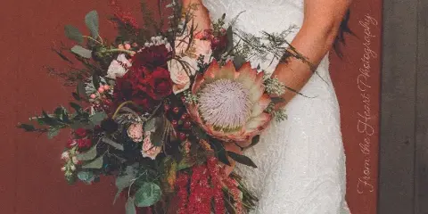 A bride in a white lace gown holding a large, custom wedding bouquet with a central protea, red roses, and trailing greenery.