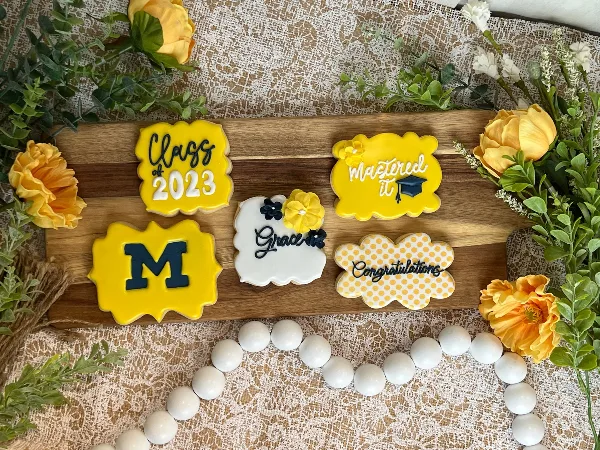 A wooden board displaying five custom-decorated sugar cookies with graduation themes, including 'Class of 2023', 'Mastered it', and 'Congratulations', surrounded by yellow flowers and greenery.