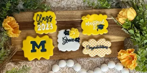 A wooden board displaying five custom-decorated sugar cookies with graduation themes, including 'Class of 2023', 'Mastered it', and 'Congratulations', surrounded by yellow flowers and greenery.