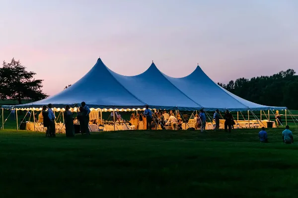 A large white event tent with a multi-peak roof set up on a grassy field at dusk, with guests and tables visible inside.
