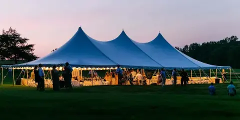 A large white event tent with a multi-peak roof set up on a grassy field at dusk, with guests and tables visible inside.
