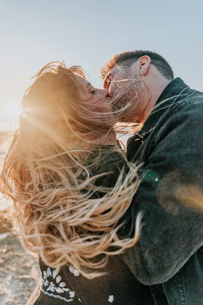 A candid, natural-light photograph of a couple kissing outdoors with wind-blown hair.
