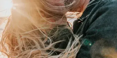 A candid, natural-light photograph of a couple kissing outdoors with wind-blown hair.