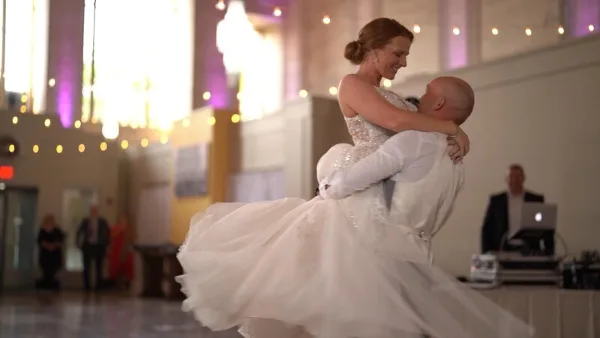 A bride and groom dancing together at a wedding reception with string lights and a chandelier in the background.