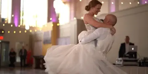 A bride and groom dancing together at a wedding reception with string lights and a chandelier in the background.