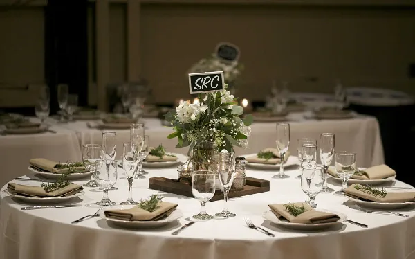 A round event table set with white linens, plates, glassware, flatware, and a floral centerpiece, with an 'SRC' sign.