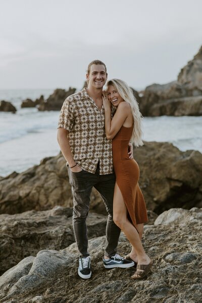 A smiling couple posing together on a rocky beach with the ocean in the background.