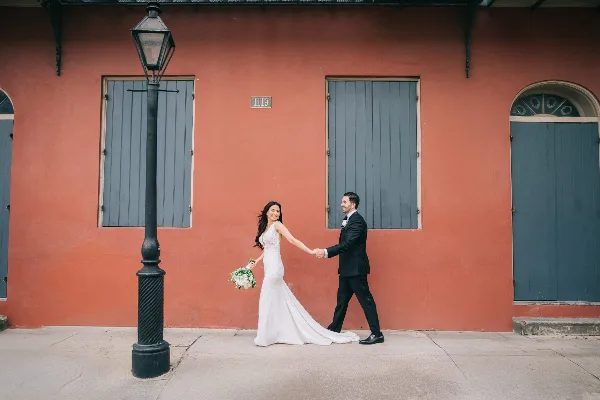 A bride and groom walking hand-in-hand on a sidewalk in front of a terracotta-colored building with blue shutters and a vintage street lamp.