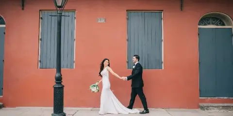 A bride and groom walking hand-in-hand on a sidewalk in front of a terracotta-colored building with blue shutters and a vintage street lamp.
