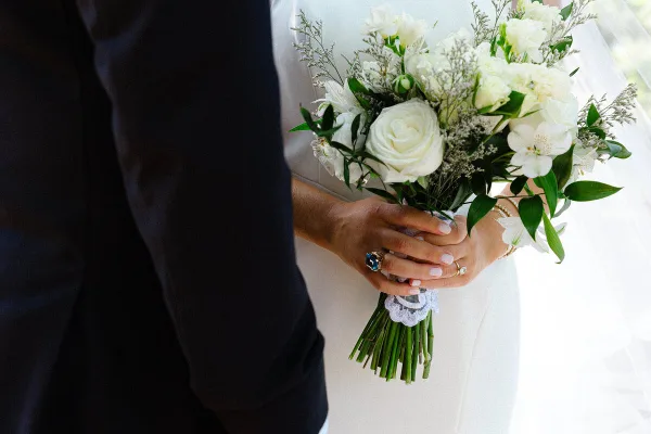 A close-up photograph of a bride's hands holding a white floral bouquet, showing wedding rings and a lace detail on the dress.