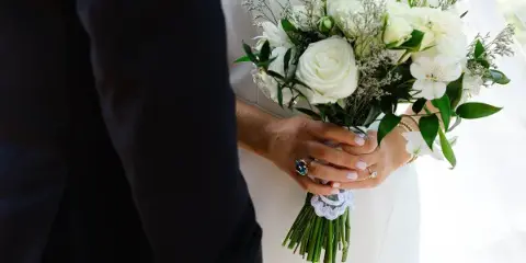 A close-up photograph of a bride's hands holding a white floral bouquet, showing wedding rings and a lace detail on the dress.