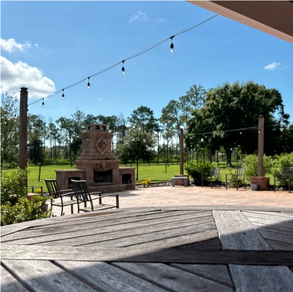 Outdoor patio area at a wedding venue featuring a stone fireplace, wooden chairs, and string lights under a blue sky.