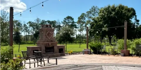 Outdoor patio area at a wedding venue featuring a stone fireplace, wooden chairs, and string lights under a blue sky.