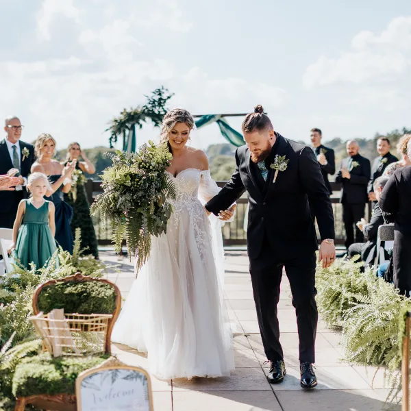 A bride and groom walking down the aisle during an outdoor wedding ceremony with guests and floral decorations.