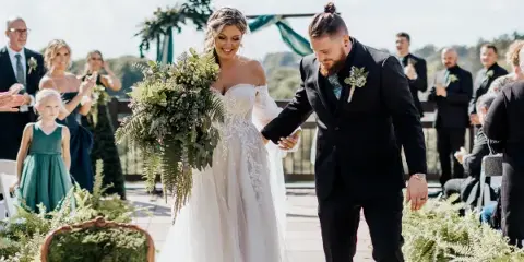 A bride and groom walking down the aisle during an outdoor wedding ceremony with guests and floral decorations.