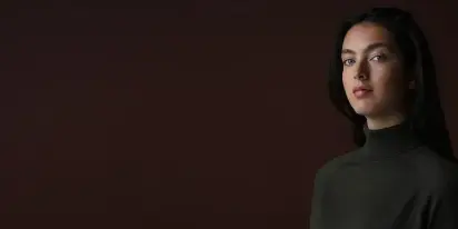 Cinematic portrait of a woman with dark hair wearing a dark turtleneck against a dark background.