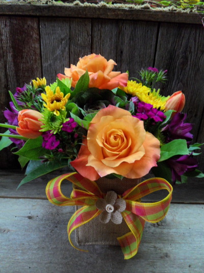 A floral arrangement featuring orange silk roses, yellow and purple flowers, and a plaid ribbon bow in a burlap-wrapped container.