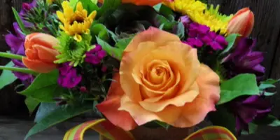 A floral arrangement featuring orange silk roses, yellow and purple flowers, and a plaid ribbon bow in a burlap-wrapped container.