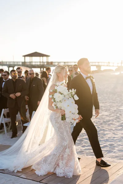 Bride and groom walking on a beach during a wedding ceremony with guests in the background.