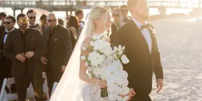 Bride and groom walking on a beach during a wedding ceremony with guests in the background.