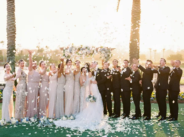 A wedding party including the bride, groom, and wedding party celebrating with flower petals outdoors.