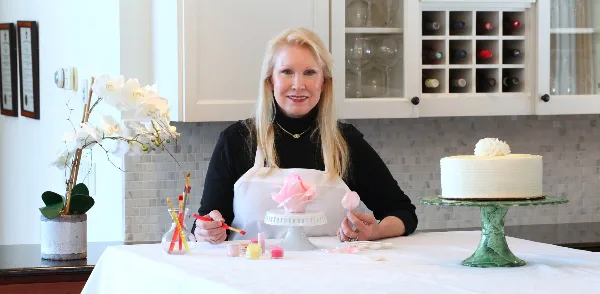 A woman in an apron sits at a workbench painting a pink sugar paste flower, with a white wedding cake on a stand to her right.