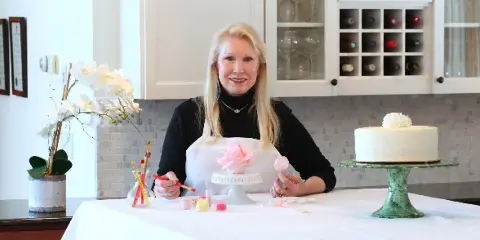 A woman in an apron sits at a workbench painting a pink sugar paste flower, with a white wedding cake on a stand to her right.