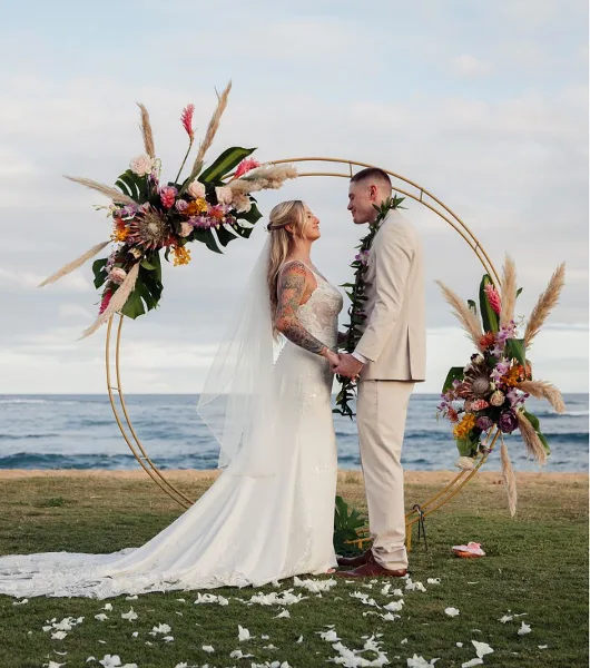 A bride and groom standing under a floral arch on a beach during a wedding ceremony.
