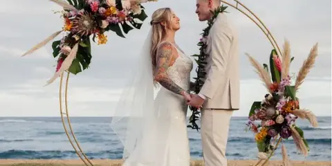 A bride and groom standing under a floral arch on a beach during a wedding ceremony.