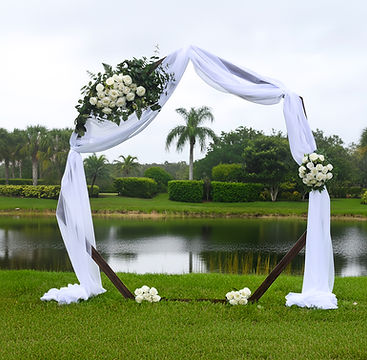 A wooden wedding arbor draped with white fabric and decorated with white flowers and greenery, set on a lawn near a body of water.