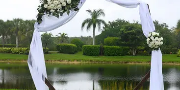 A wooden wedding arbor draped with white fabric and decorated with white flowers and greenery, set on a lawn near a body of water.