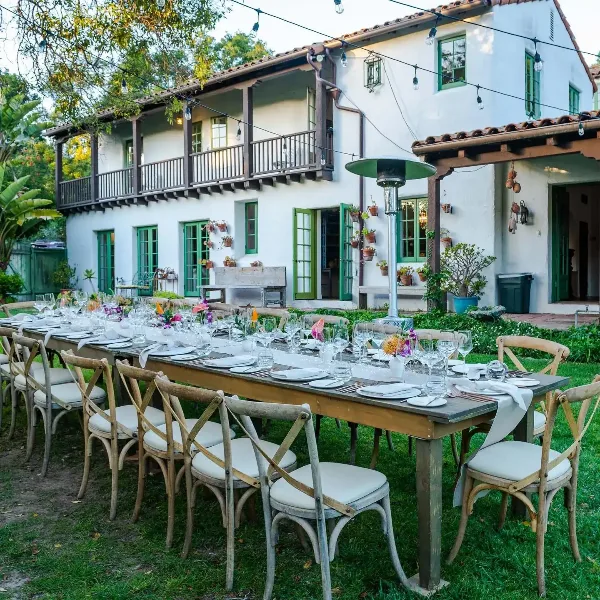 Outdoor event table set with white plates, glassware, and floral centerpieces in front of a white building with green shutters.