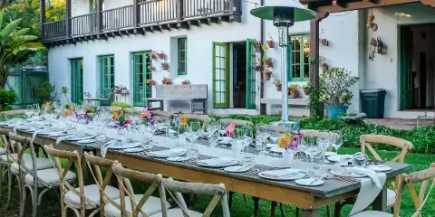 Outdoor event table set with white plates, glassware, and floral centerpieces in front of a white building with green shutters.