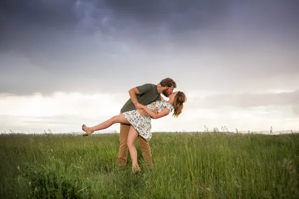 A couple embracing and kissing in a grassy field under a dramatic cloudy sky.