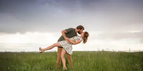 A couple embracing and kissing in a grassy field under a dramatic cloudy sky.