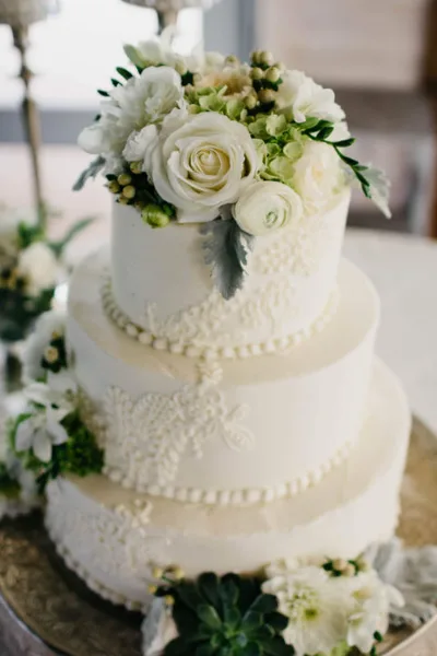 A three-tiered white wedding cake with piped lace details and a floral arrangement on top.
