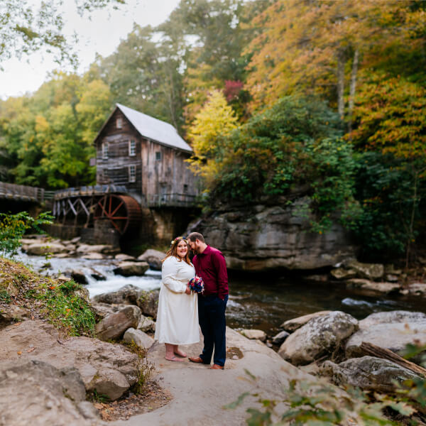 A bride and groom standing together on a rocky path in front of a historic wooden water mill surrounded by autumn foliage.