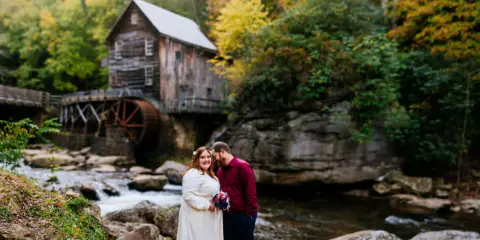 A bride and groom standing together on a rocky path in front of a historic wooden water mill surrounded by autumn foliage.