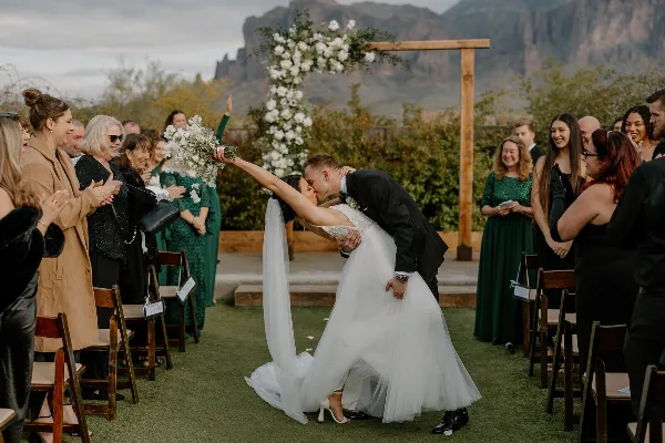 A bride and groom share a dip kiss at the altar during an outdoor wedding ceremony, surrounded by cheering guests and a floral arch.