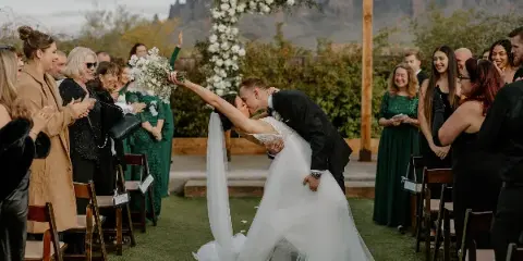 A bride and groom share a dip kiss at the altar during an outdoor wedding ceremony, surrounded by cheering guests and a floral arch.