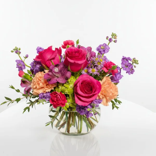 A mixed floral bouquet with pink roses, purple alstroemeria, and peach carnations in a round glass vase.