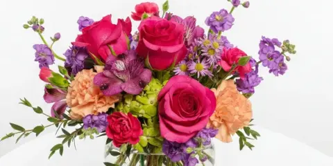 A mixed floral bouquet with pink roses, purple alstroemeria, and peach carnations in a round glass vase.