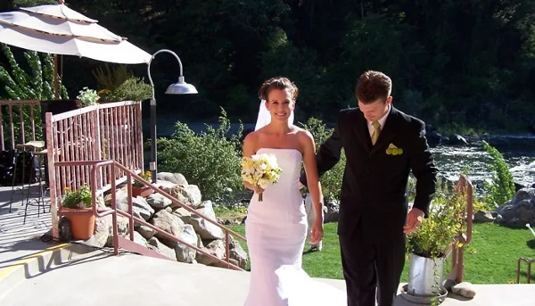 A bride and groom walking on a patio with a river and greenery in the background.