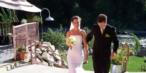 A bride and groom walking on a patio with a river and greenery in the background.