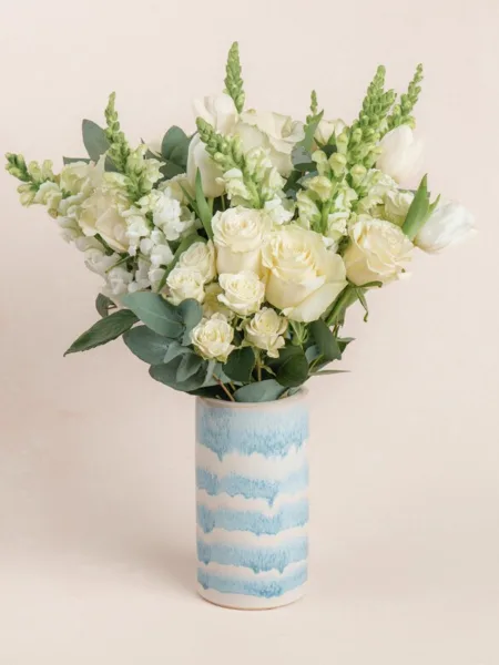 A white floral bouquet containing roses, snapdragons, and eucalyptus arranged in a blue and white patterned ceramic vase.