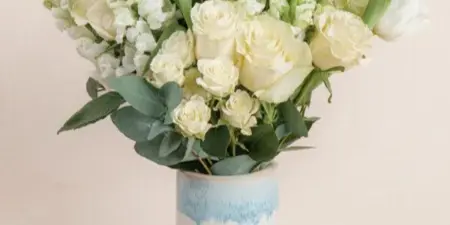 A white floral bouquet containing roses, snapdragons, and eucalyptus arranged in a blue and white patterned ceramic vase.
