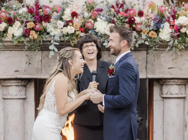 A professional wedding officiant presiding over a ceremony between a bride and groom in front of a floral arch.